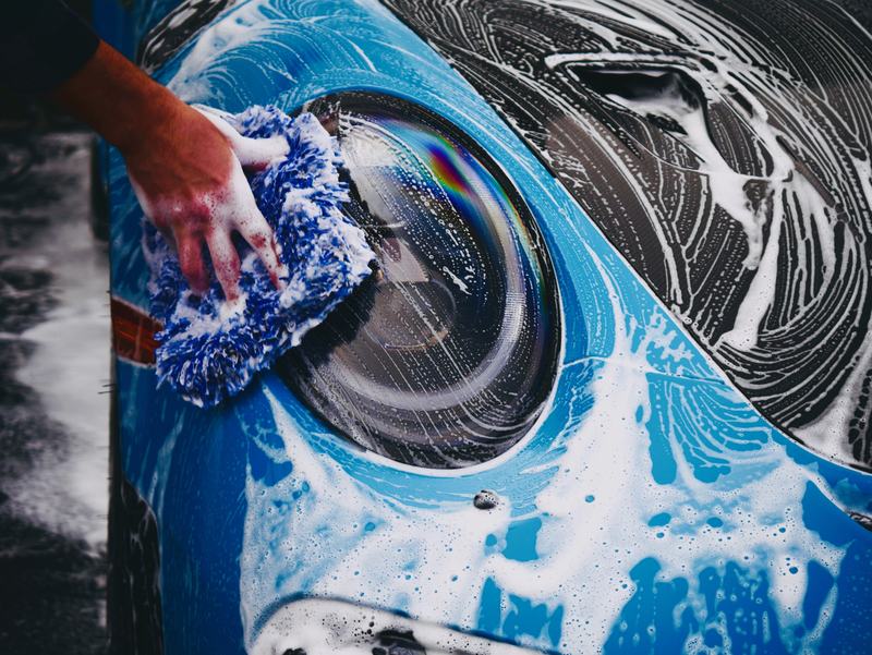 A hand washing a blue Porsche headlight with a wash mitt and thick soap foam